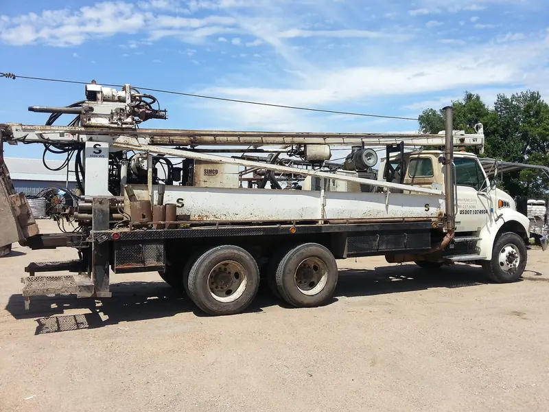 A truck-mounted water well drilling rig with its mast folded along the bed, seen from the side in a gravel yard.
