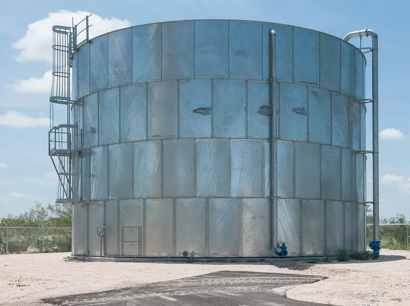 A tall galvanized steel ground storage tank with a ladder and inlet piping, standing on a gravel pad in South Texas.